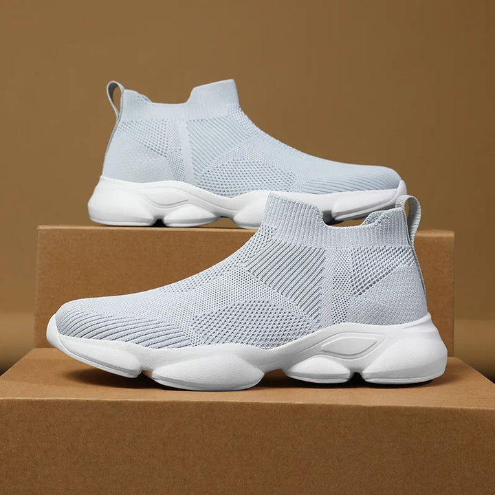 Light gray slip-on athletic sneakers made with lightweight, breathable knit fabric and thick white non-slip soles, displayed on brown cardboard boxes against a plain tan background.