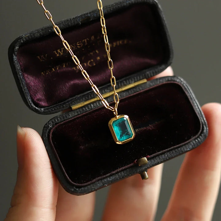 A hand holds an open black velvet jewelry box displaying an 18K gold plated necklace with a rectangular green zirconia pendant inside. The box interior is lined with dark purple fabric.