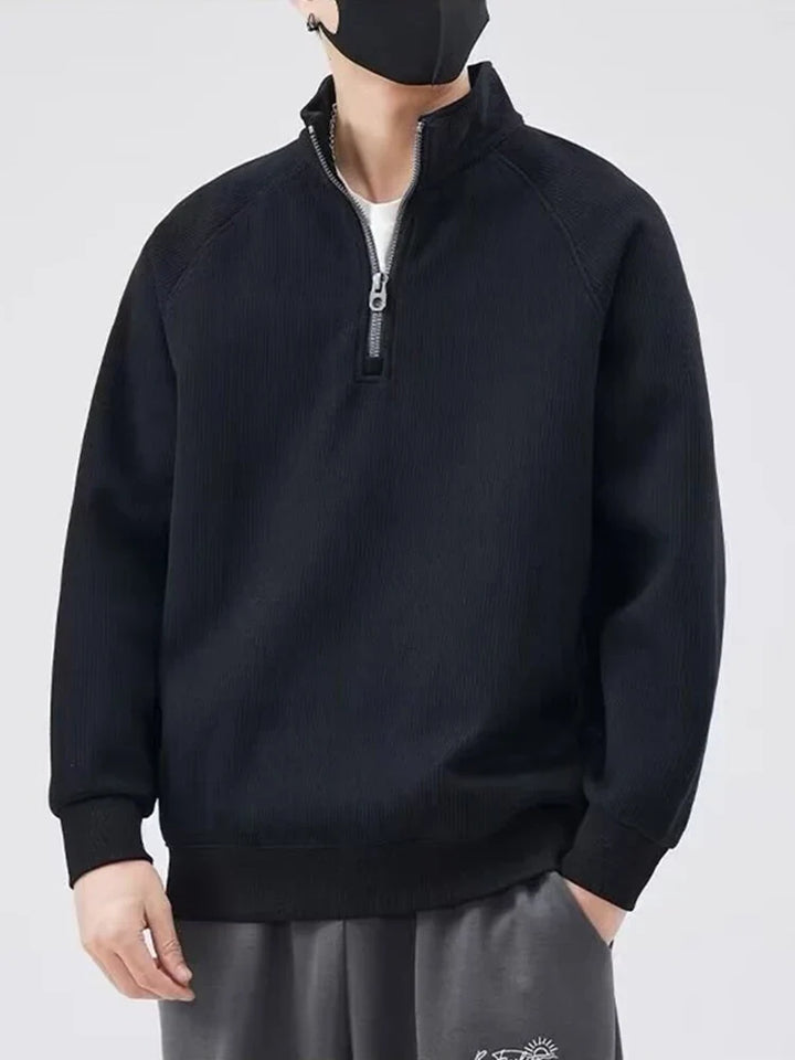 A person wearing a black zip-up pullover with a stand-up collar, gray pants, and a black face mask is standing with hands relaxed at their sides against a plain background.