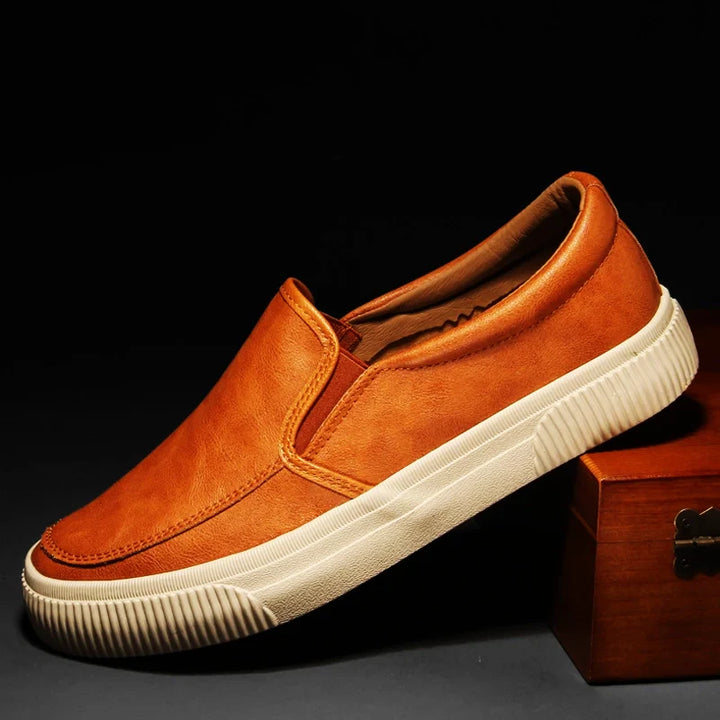 A tan men’s slip-on casual shoe with a white hybrid rubber sole is displayed against a dark background, resting with the heel elevated on a wooden box.