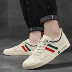 A person wearing cream-colored sneakers with green and red stripes and a breathable mesh upper, paired with rolled-up dark denim jeans, standing on a gray floor.