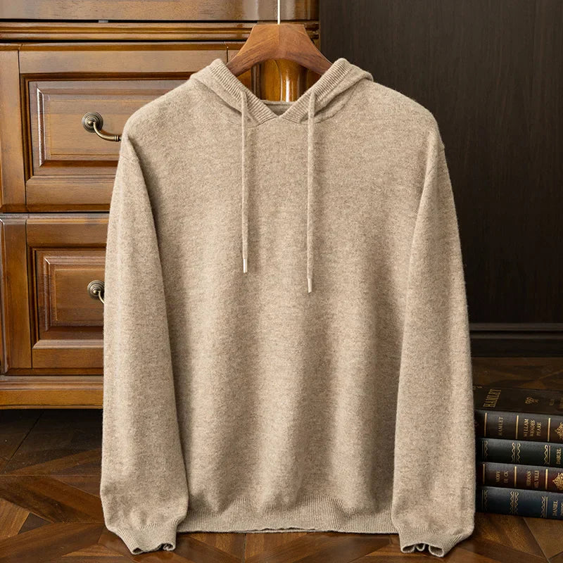 A beige cashmere blend hooded sweatshirt with drawstrings hangs on a wooden hanger, placed in front of a wooden dresser and next to a stack of books on a wooden floor. Perfect as a cozy layering sweater for chilly days.