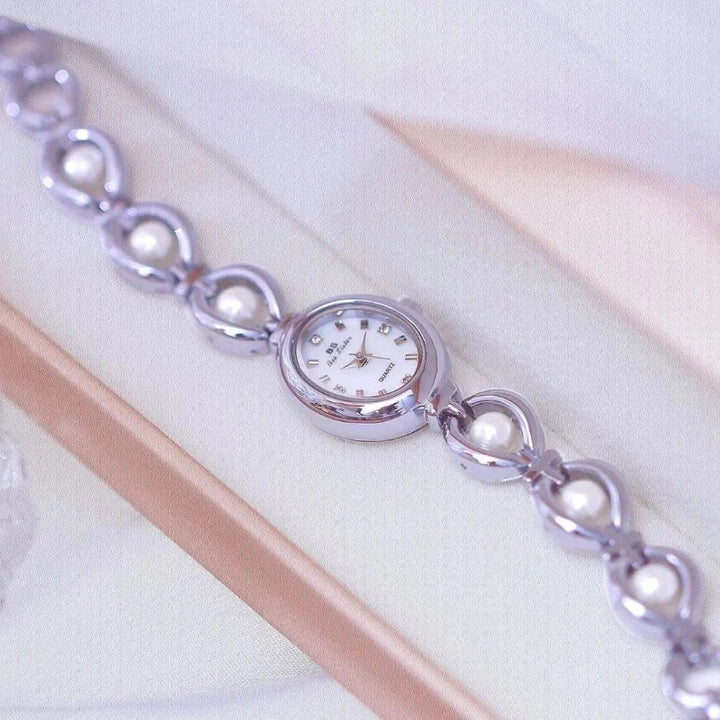 A delicate silver wristwatch with a white oval face, pearl accents along the band, and precision quartz movement, displayed on a soft, light background.