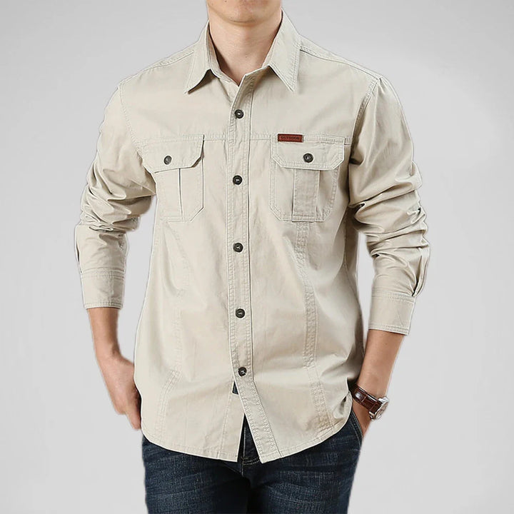 A person wearing a light beige, relaxed fit 100% cotton shirt with two chest pockets and dark buttons, paired with dark blue jeans. The person’s hands are in their pockets against a plain gray background.