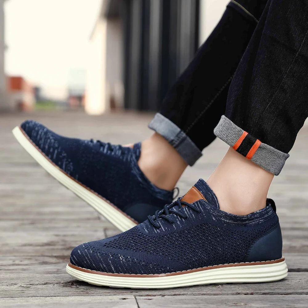 A person wearing dark blue breathable knit sneakers with white soles and rolled-up jeans stands on a wooden surface outdoors, showcasing a relaxed casual style.