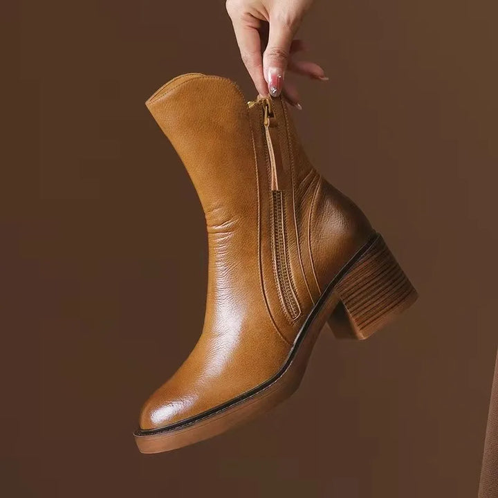 A hand holds a tan leather women's ankle boot with a chunky heel, side zipper, and rounded toe against a brown background—perfect for elevating your winter boots collection.