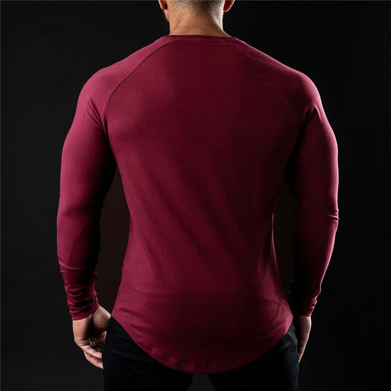 A man with short hair is shown from behind, wearing a long sleeve fitted shirt in burgundy and black pants, standing against a dark background.