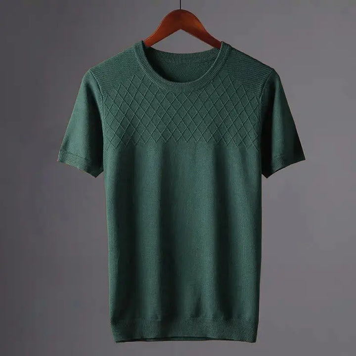 A green short-sleeve knit sweater in a classic fit hangs on a wooden hanger, featuring a diamond-pattern across the chest and a plain lower section, set against a neutral gray background.