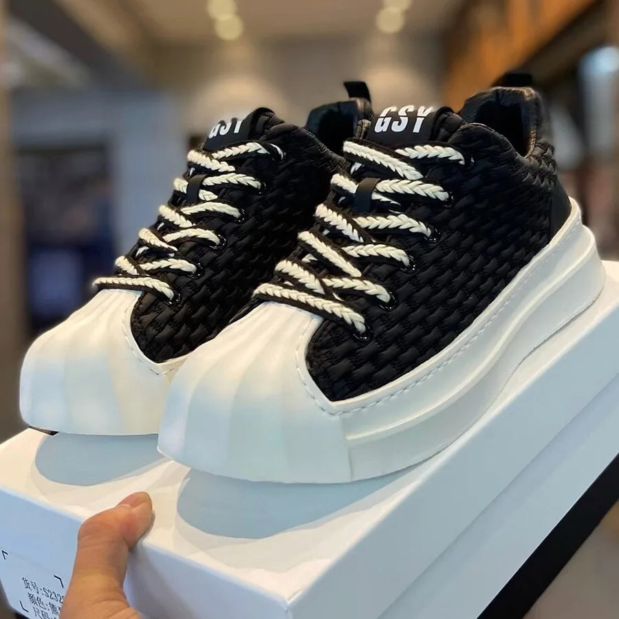 A hand holds a box with black woven upper sneakers featuring thick cushioned soles, rounded toe caps, white and black rope laces, and “GSY” printed on the tongue, displayed in a store setting.