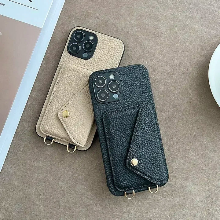 Two smartphones in beige and black pebble-texture material cases with attached cardholder pockets and gold button clasps rest on a soft brown surface beside a white saucer and a partially visible book or magazine.