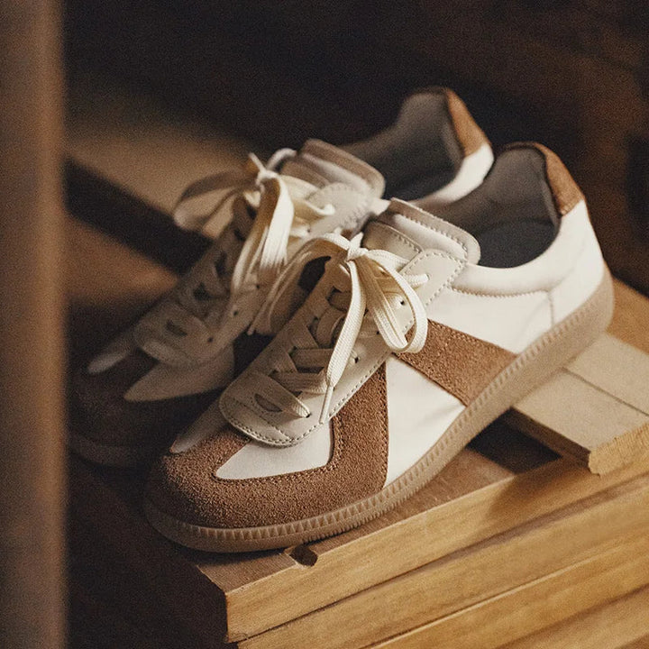 A pair of white and brown premium cow suede sneakers with cream laces, featuring lightweight breathable design, placed on a wooden surface in soft, warm lighting.