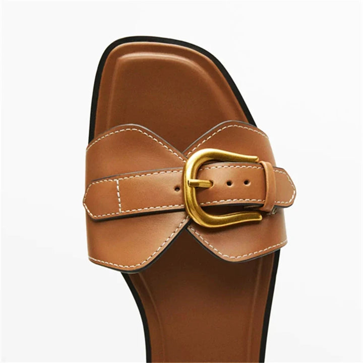 A close-up of a tan leather slip-on sandal with white stitching, featuring a large gold buckle on the strap, set against a plain white background.