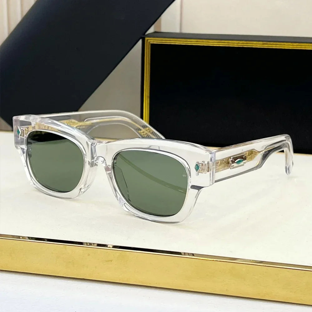 Vintage-inspired sunglasses with clear frames and green-tinted lenses rest on a white and gold surface, while a black and gold box sits in the background.