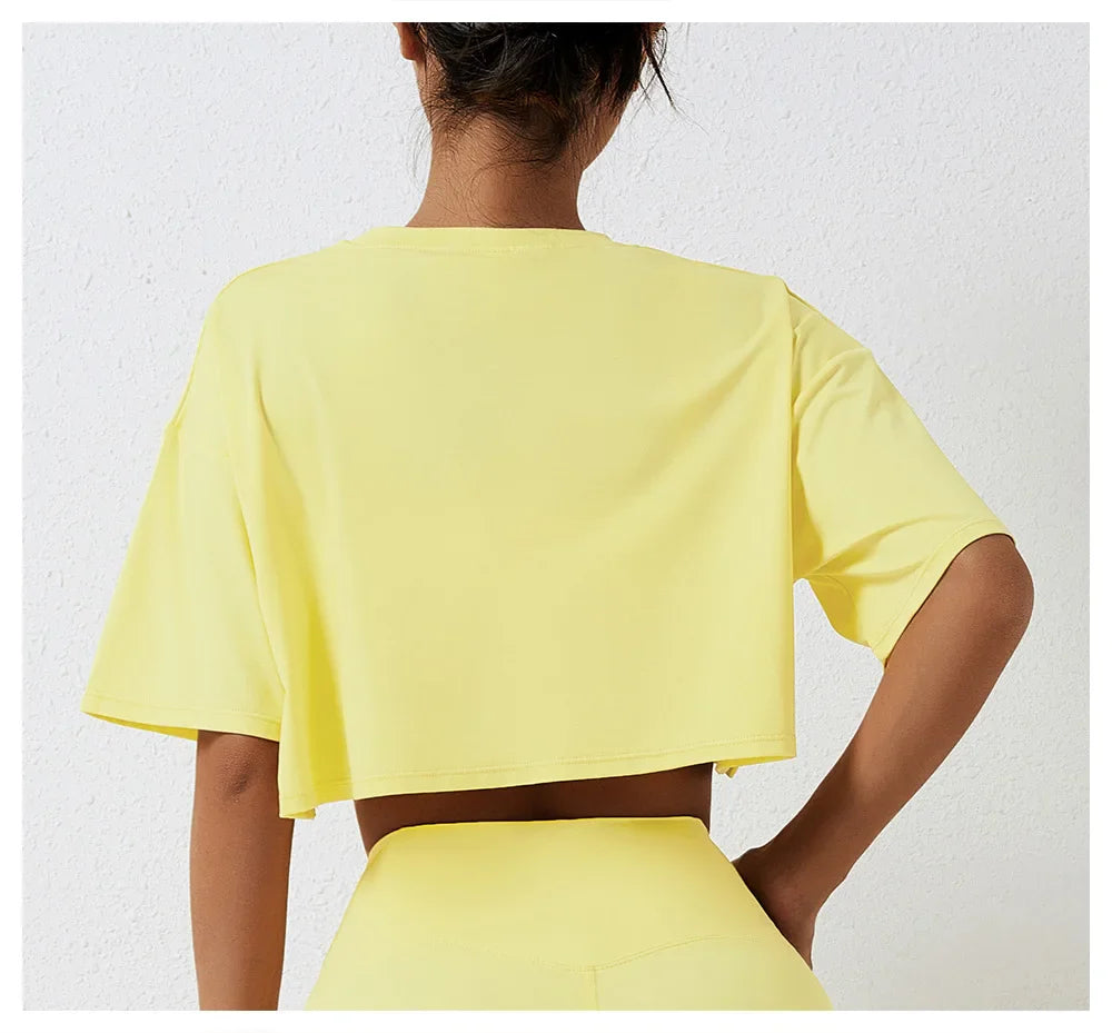 A person stands facing away from the camera, wearing a relaxed fit, short-sleeved yellow cotton crop top and matching high-rise pants, set against a white textured wall.