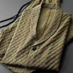 A folded olive green textured jacket with a shawl collar and single button, placed next to matching pants with an elastic drawstring waistband on a dark surface. The fabric features a raised checkered pattern.