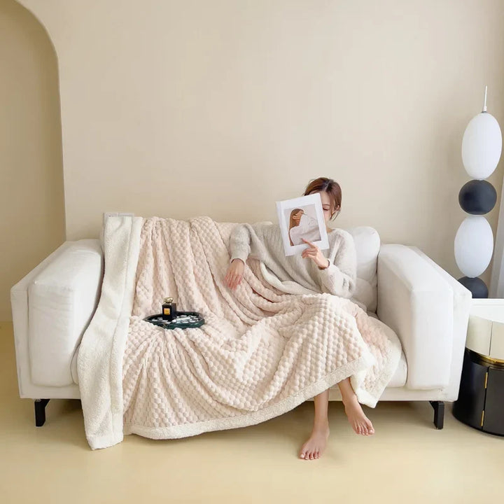 A person sits on a white sofa, wrapped in an ultra-soft cotton blanket, reading a magazine. A tray with a candle and drink rests nearby. The room is minimal with soft lighting and neutral colors.