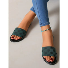 A person wearing blue skinny jeans, green open-toe slide silhouette sandals with a subtle black pattern, and a turquoise beaded anklet, resting their feet on a light-colored floor.