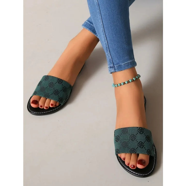 A person wearing blue skinny jeans, green open-toe slide silhouette sandals with a subtle black pattern, and a turquoise beaded anklet, resting their feet on a light-colored floor.