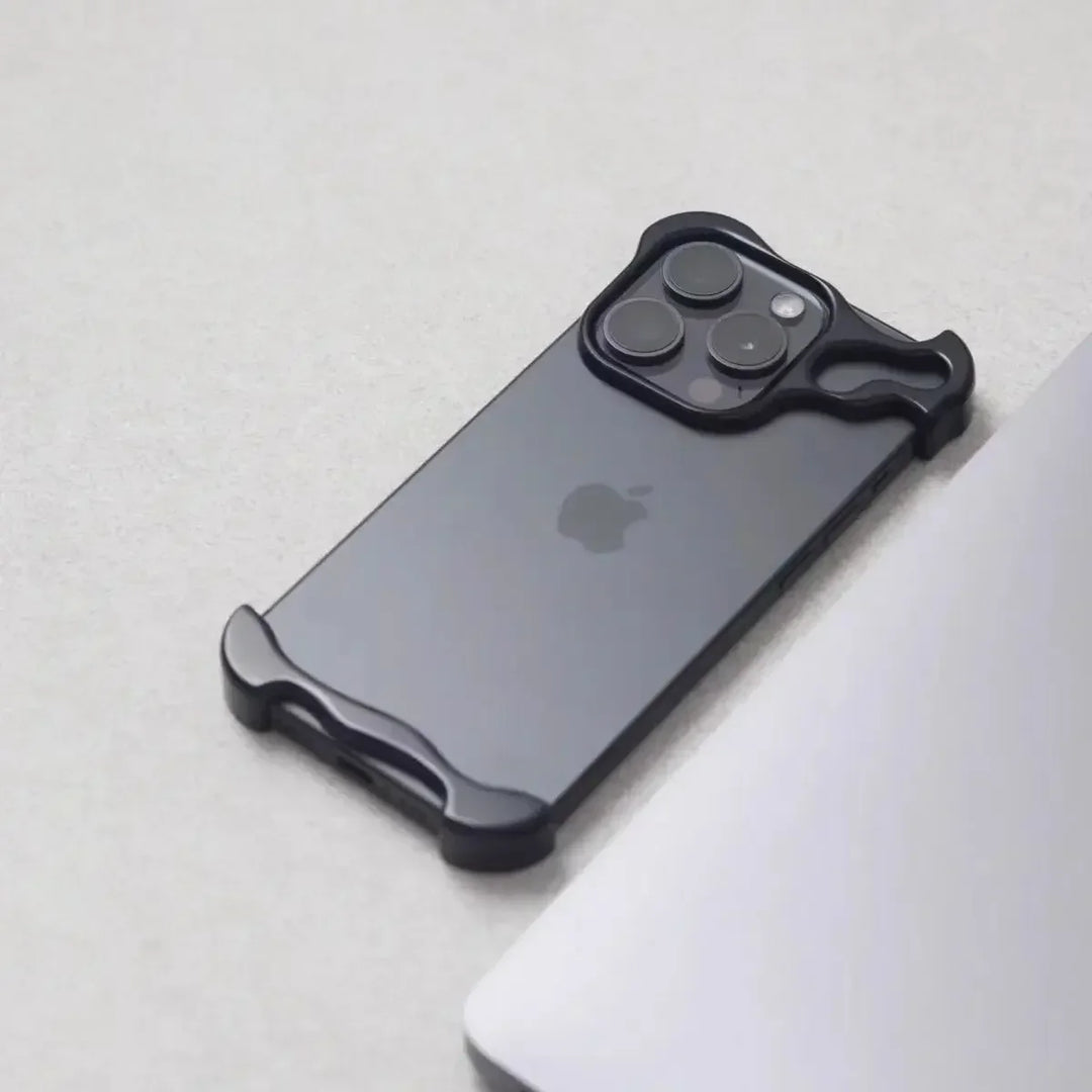 A black iPhone with a minimalist, Metal Bumper Design case is placed on a light gray surface next to the edge of a silver laptop. The case covers only the corners and camera area of the phone.
