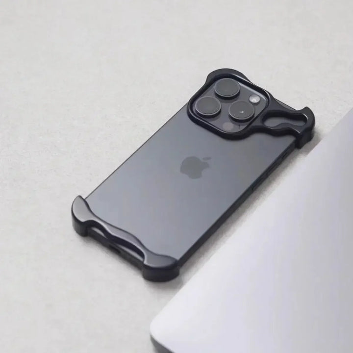 A black iPhone with a minimalist, Metal Bumper Design case is placed on a light gray surface next to the edge of a silver laptop. The case covers only the corners and camera area of the phone.