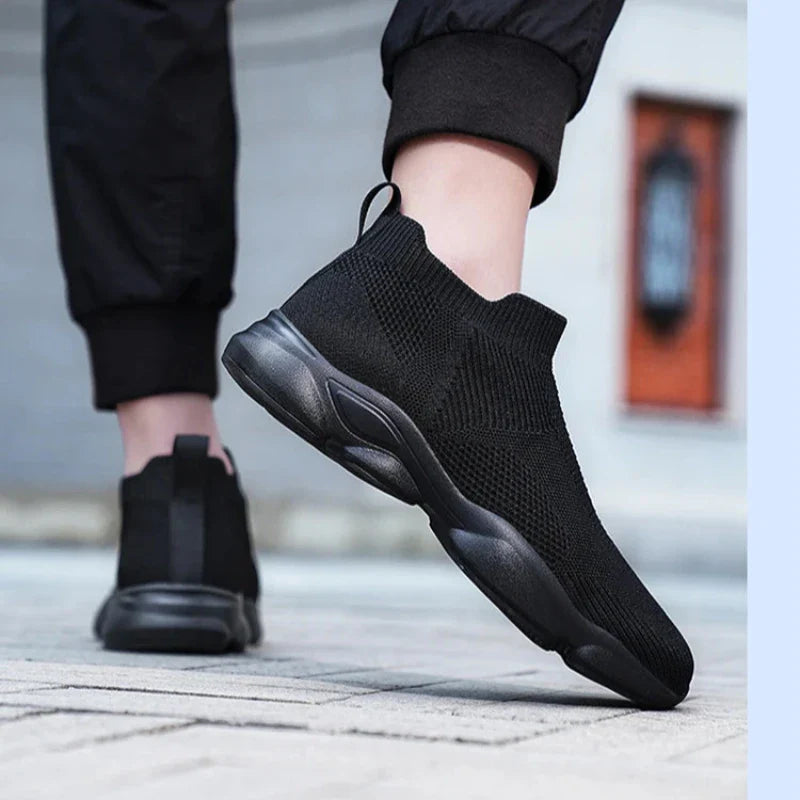 A person wearing black slip-on athletic sneakers with a non-slip sole and black pants stands on a paved surface, lifting one foot slightly off the ground. The background is blurred, keeping the focus on the shoes.