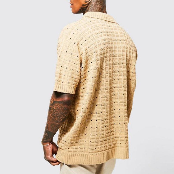 A person with tattooed arms is wearing a loose-fitting, short-sleeved summer shirt in beige, featuring a breathable knit design with a textured square pattern, viewed from the back against a plain light gray background.