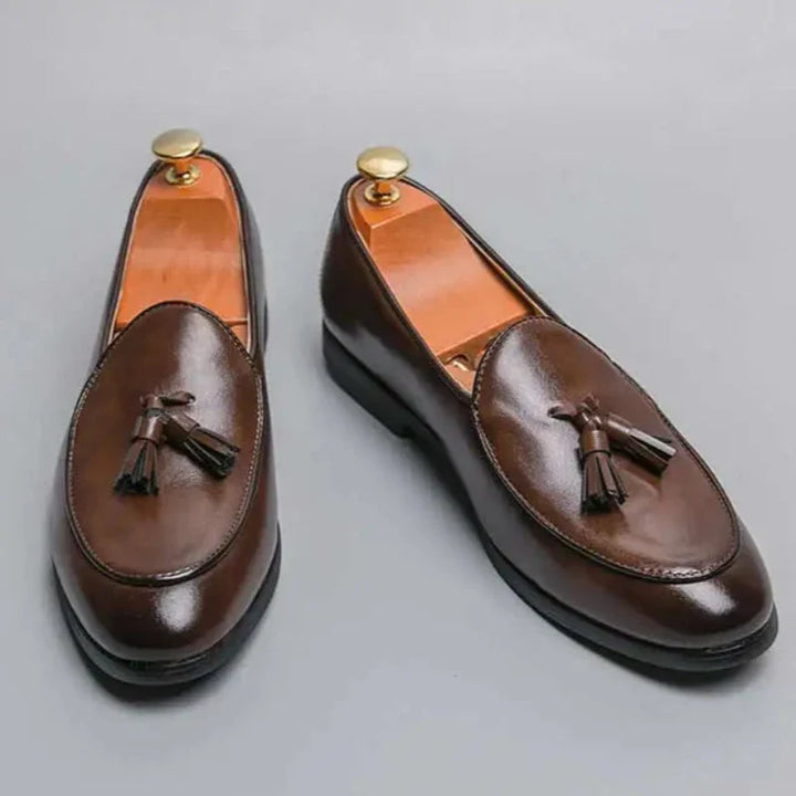 A pair of polished brown leather loafers with tassels on top, each fitted with a gold shoe tree, displayed on a light gray background for a refined vintage look.
