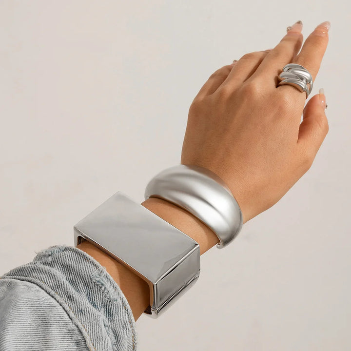 A hand wearing chunky silver jewelry—geometric design pieces like a thick rectangular bracelet, a wide rounded bangle, and a sculptural silver ring. The person is dressed in light grey textured fabric, showcasing bold unisex jewelry.