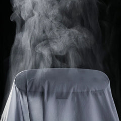 A close-up of a lightweight white shirt draped over a surface with steam rising from it against a black background, suggesting the shirt is being ironed or steamed.