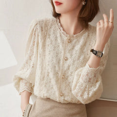 A woman wears an ivory lace blouse with vintage pearl buttons and puffed sleeves, paired with a beige skirt. She accessorizes with a watch, ring, earrings, and has shoulder-length brown hair.