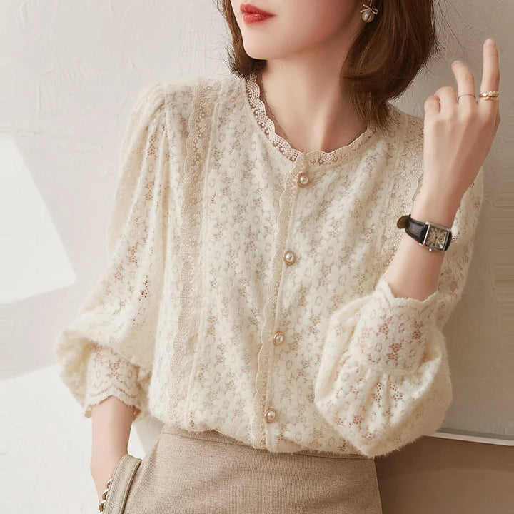 A woman wears an ivory lace blouse with vintage pearl buttons and puffed sleeves, paired with a beige skirt. She accessorizes with a watch, ring, earrings, and has shoulder-length brown hair.