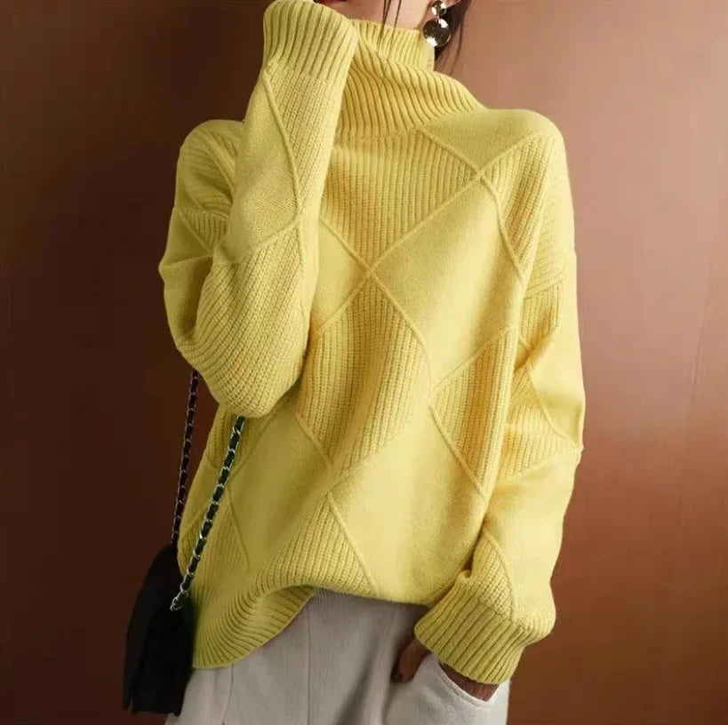 A person wearing a light yellow, diamond knit turtleneck sweater with a geometric pattern, one hand near their face, paired with light-colored pants and a black shoulder bag.