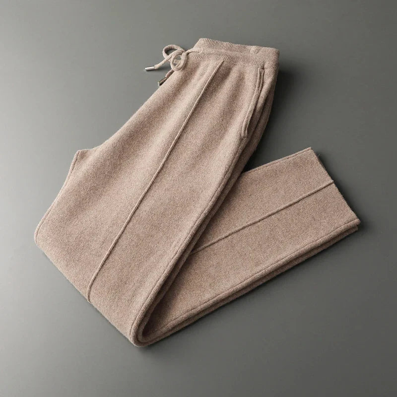 A pair of neatly folded beige knit pants in a tailored pencil cut with an elastic waistband and drawstring, crafted from a soft cashmere wool blend, lying on a plain gray surface.