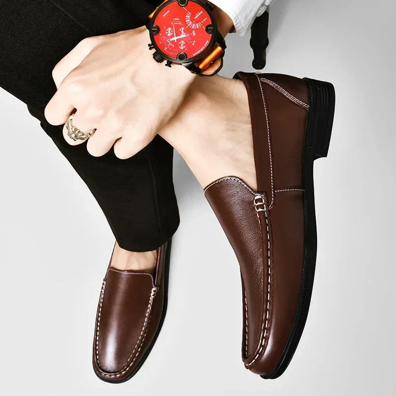 A person wearing classic slip-on genuine leather loafers and black pants rests one leg over the other, showing a wrist with a large red-faced watch and a gold ring. The background is plain and light-colored.