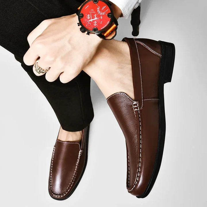 A person wearing classic slip-on genuine leather loafers and black pants rests one leg over the other, showing a wrist with a large red-faced watch and a gold ring. The background is plain and light-colored.