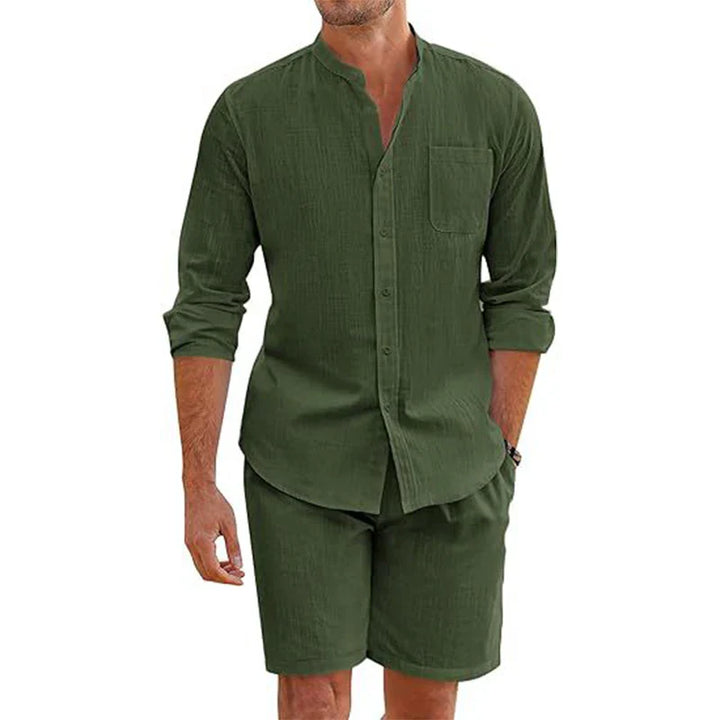 A man wears a matching dark green cotton linen outfit: a vintage stand collar shirt with chest pocket and rolled-up sleeves, paired with baggy shorts that fall to his knees. He stands relaxed, one hand by his side.
