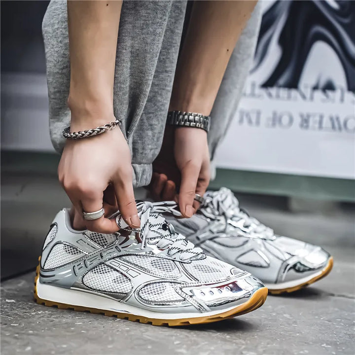 A person wearing gray sweatpants and lightweight mesh shoes with a cushioned insole is crouching down and tying their laces. Their hands display a watch, bracelet, and rings.