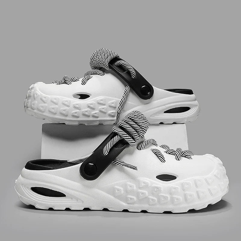 A pair of white slip-on clogs with textured soles for enhanced grip, black interiors, and black-and-white striped lace detail and straps, displayed against a gray background with one shoe propped on a white block.