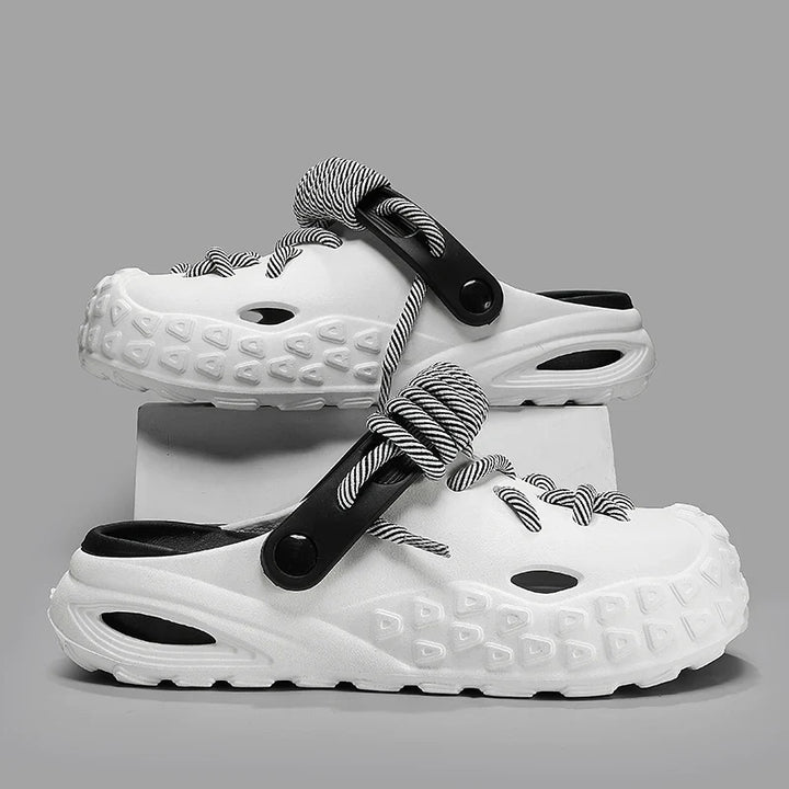 A pair of white slip-on clogs with textured soles for enhanced grip, black interiors, and black-and-white striped lace detail and straps, displayed against a gray background with one shoe propped on a white block.