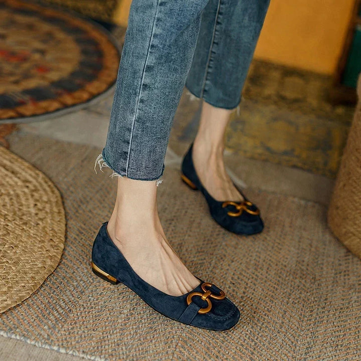 A person wearing cropped, faded blue jeans and navy blue suede flats with a cushioned insole and gold decorative buckles stands on a woven rug.