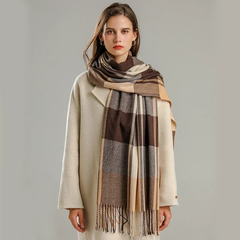 A woman with long brown hair wears a cream-colored coat and a large, lightweight plaid scarf in shades of brown, beige, and gray. She stands against a plain, light gray background, looking at the camera.