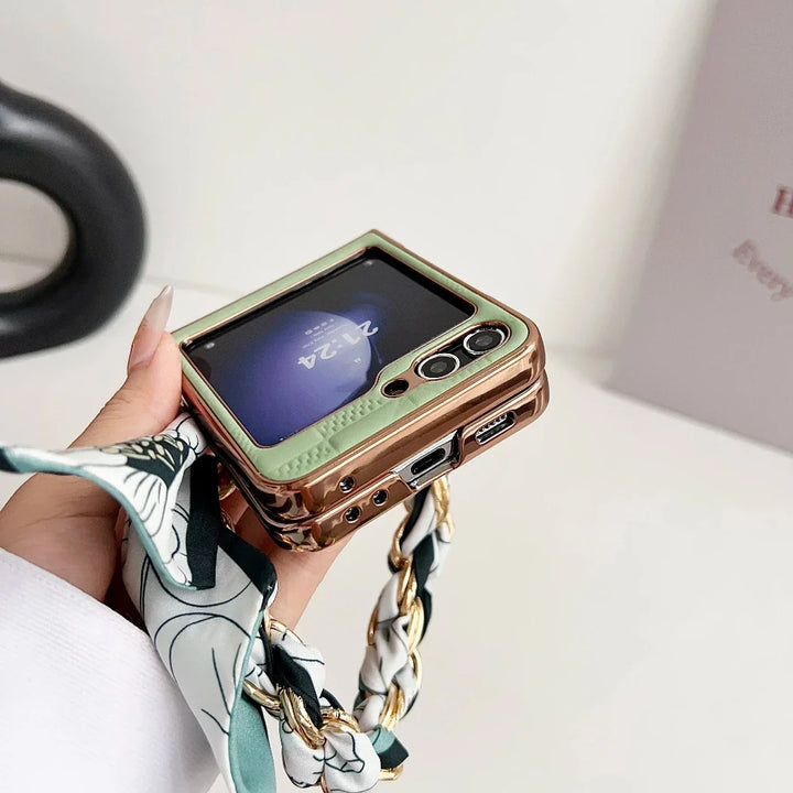 A hand holds a gold and green Samsung Galaxy Z Flip with a visible screen displaying the time. The phone features a premium leather case, a decorative strap with patterned fabric and chain. The background is minimal and out of focus.