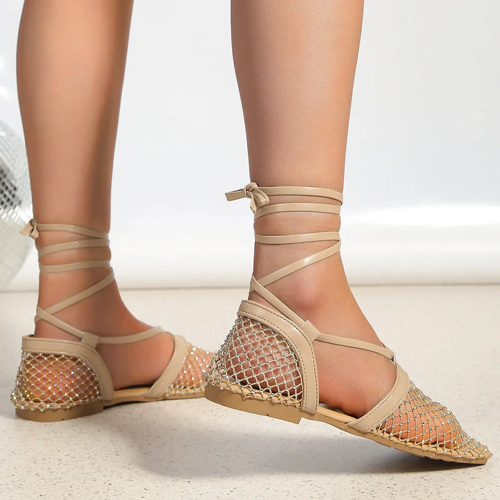 A person wearing beige flat sandals with a mesh upper and ankle wrap straps, featuring slip-resistant soles, stands on a light-colored floor.