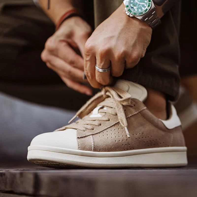 A person wearing a watch and ring crouches to tie the laces of a beige and white minimalist sneaker, crafted from premium canvas with perforated sides and a white sole.