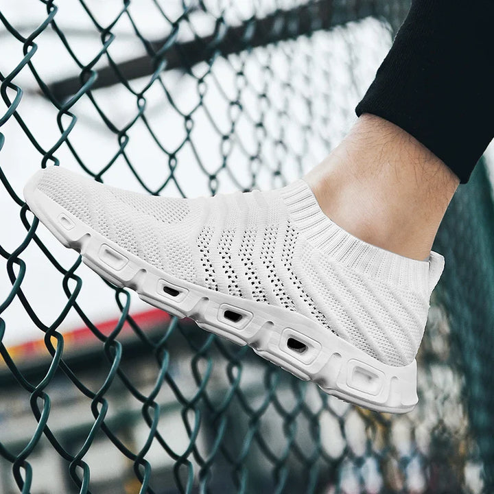 A person wearing a white, knit slip-on sneaker with a sock-like fit and thick, textured sole stands near a metal chain-link fence. The shoe features breathable material and a modern, sporty design.