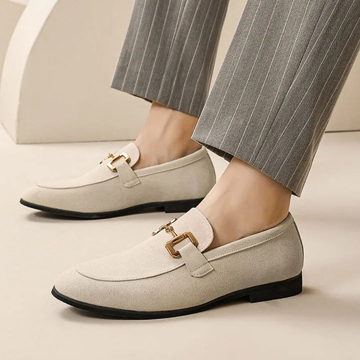 A person wearing luxury loafers with gold buckle details and gray pinstripe pants stands on a beige surface.