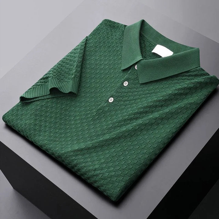 A neatly folded dark green polo shirt with a textured, waffle jacquard diamond-patterned fabric, three buttons, and a classic collar, displayed on a black and gray surface.