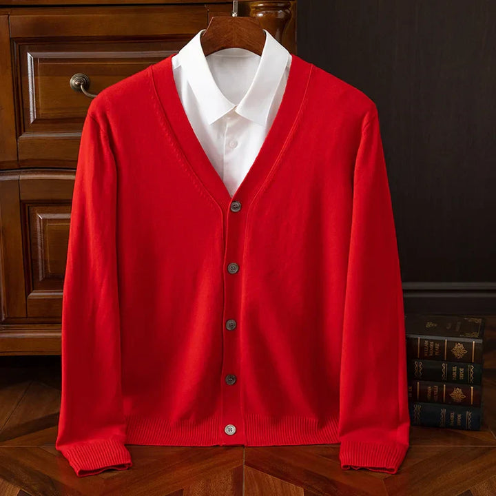 A bright red, V-neckline button-up cardigan in a relaxed fit is displayed on a hanger over a white collared shirt, set against a wooden dresser and a stack of books on a wooden floor.
