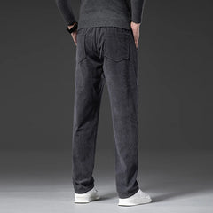 A person wearing a dark sweater, men's smart casual pants in dark gray corduroy with a zipper fly, and white sneakers stands facing away, hands behind their back, against a plain gray background.