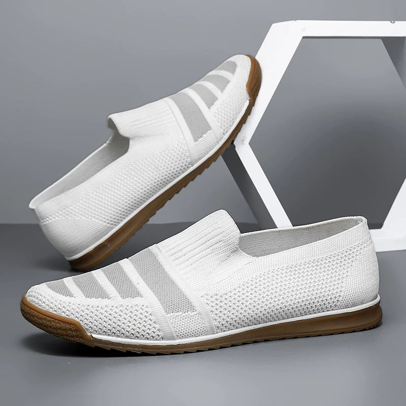 A pair of white slip-on men's casual shoes with textured knit material and brown rubber soles, offering a breathable loafers design, displayed against a gray background with a geometric white decor piece.